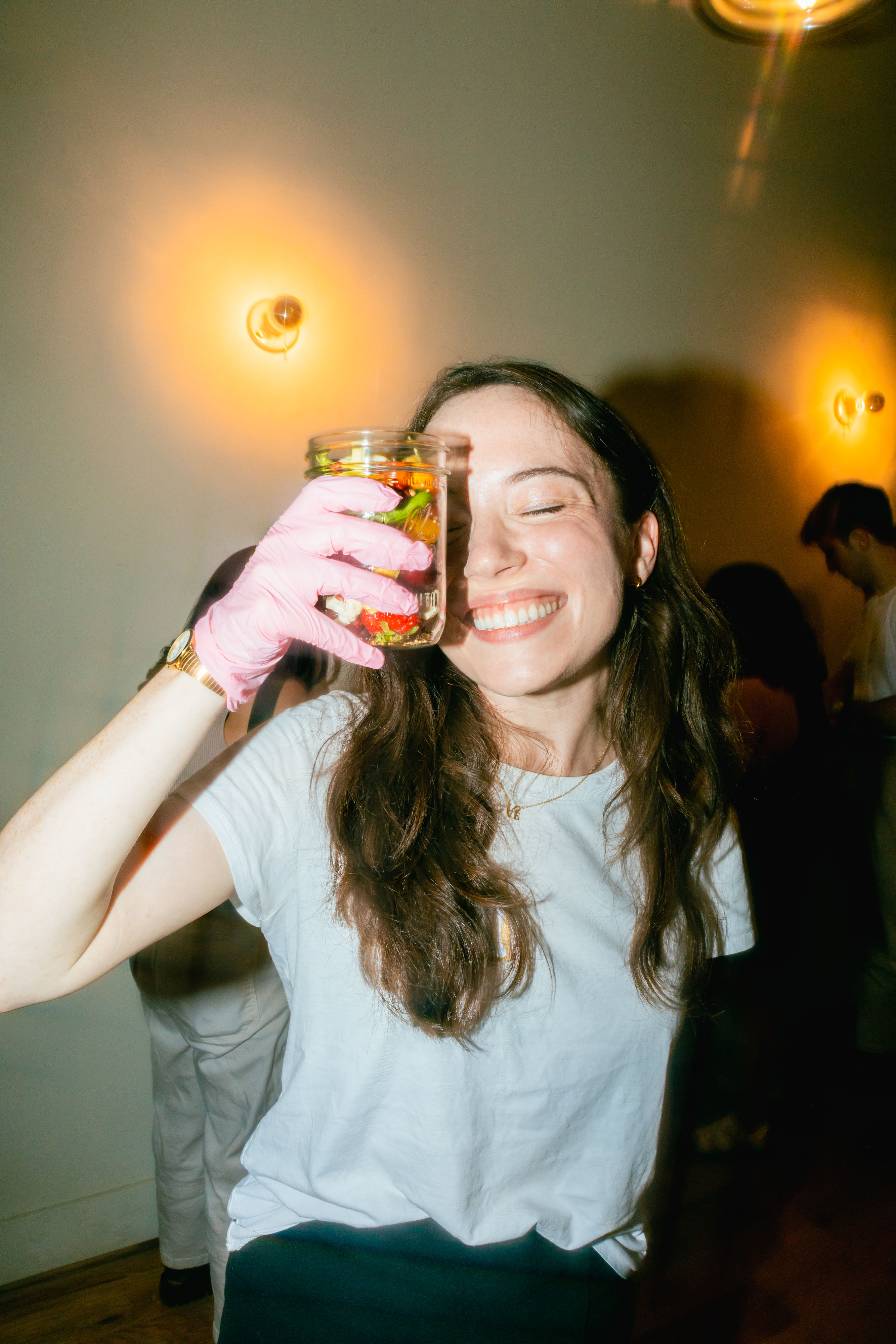 Smiling woman at an indoor party holding a mason jar of colorful pickled vegetables up to her eye while wearing a pink glove, warm wall sconces glowing in the background