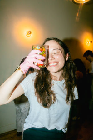 Smiling woman at an indoor party holding a mason jar of colorful pickled vegetables up to her eye while wearing a pink glove, warm wall sconces glowing in the background