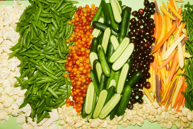 Colorful fresh produce display arranged in rows: cauliflower, sugar snap peas, orange and red cherry tomatoes, sliced cucumbers, dark grapes, rainbow carrots and peeled garlic cloves on a pale green background.