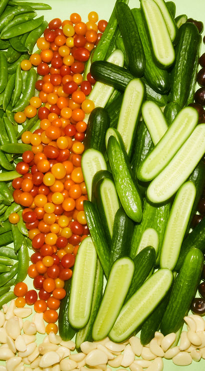 Colorful fresh produce display arranged in rows: cauliflower, sugar snap peas, orange and red cherry tomatoes, sliced cucumbers, dark grapes, rainbow carrots and peeled garlic cloves on a pale green background.