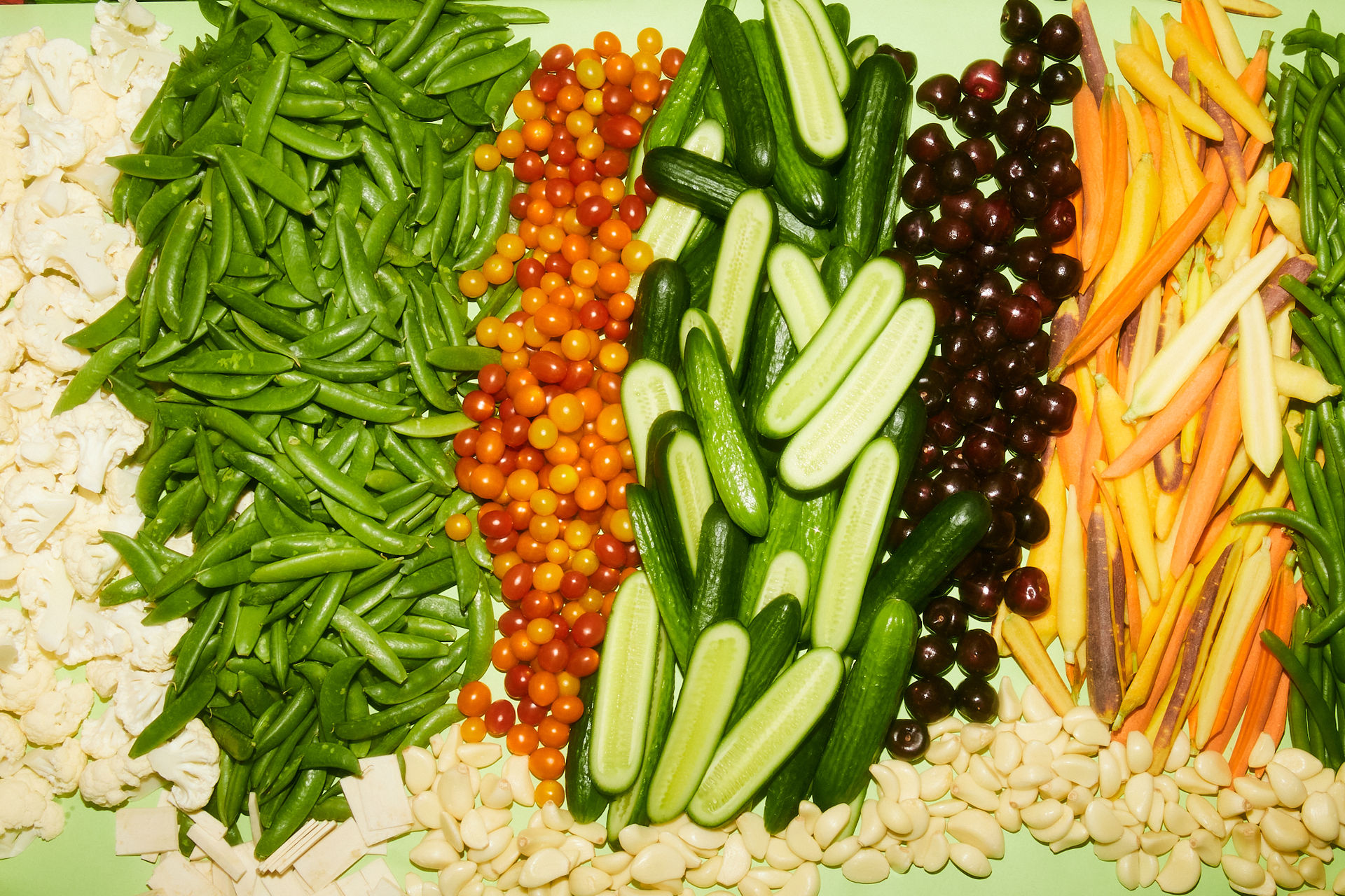 Colorful fresh produce display arranged in rows: cauliflower, sugar snap peas, orange and red cherry tomatoes, sliced cucumbers, dark grapes, rainbow carrots and peeled garlic cloves on a pale green background.