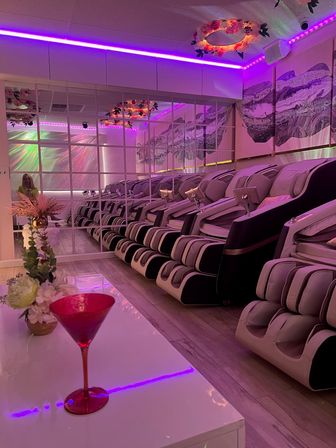 Row of reclining massage chairs in a pink‑purple LED lit spa lounge with mirrored wall, abstract wall art, floral ceiling decor and a red cocktail glass on a white table in the foreground.