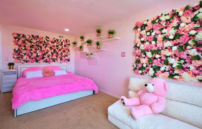 Bright pink bedroom with fluffy hot-pink bedspread, two large rose floral wall panels, floating shelves with potted plants, and a pink teddy bear on a white loveseat.