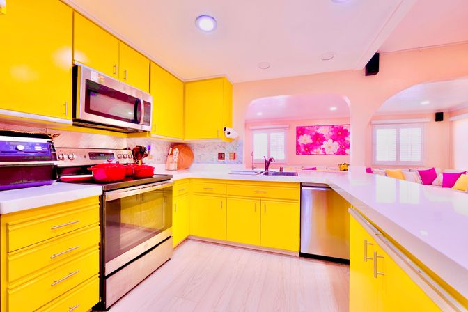 Sunny modern open-concept kitchen with glossy yellow cabinets, white quartz countertops, stainless steel stove, microwave and dishwasher, and a breakfast bar overlooking a living area with pink floral artwork and colorful throw pillows.