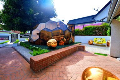 Sunlit backyard courtyard with a bronze-tinted geodesic glass dome, reflective copper spheres, brick patio, yellow decorative bench, and manicured planters beneath a leafy tree and flowering hedge.