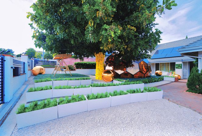 Modern suburban front yard with a large shade tree wrapped in yellow lights, tiered white planter beds of ornamental grasses, reflective gold spheres and a geometric mirrored pod, a wooden swing, gravel pathways and a house with solar panels
