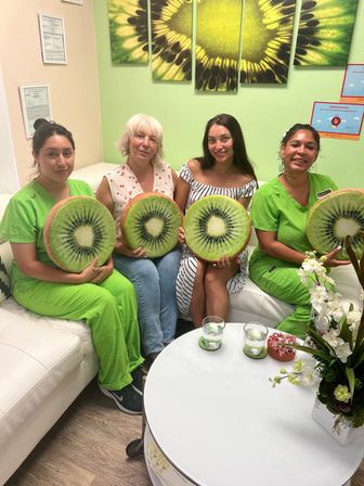 Four women seated on a white couch in a bright green-themed wellness clinic waiting room, each holding a playful kiwi-slice cushion; two wear lime scrubs, striped dress and casual top, with sunflower wall art and a white coffee table with water glasses and flowers in the foreground.