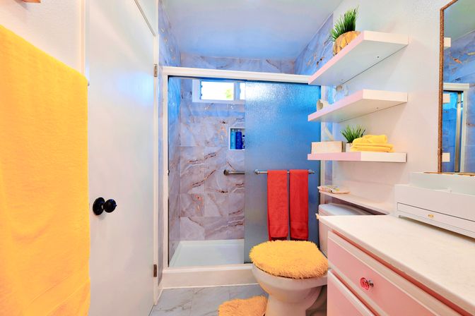 Cheerful small residential bathroom with frosted glass sliding shower, marble-look tile, pink vanity, white floating shelves, yellow towels and fuzzy toilet seat cover.