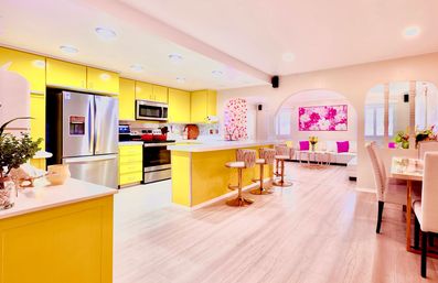 Bright yellow open-concept kitchen and living room with stainless steel appliances, long island with gold-accent bar stools, light wood floors and pink floral artwork.
