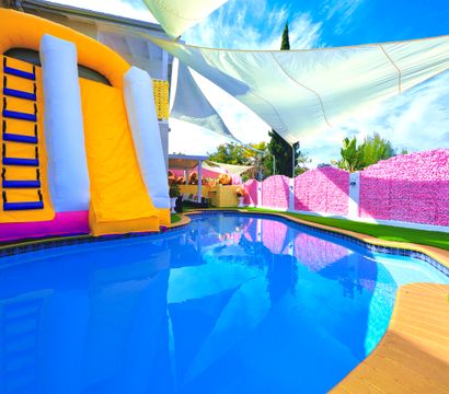 Bright backyard swimming pool with vivid blue water, a yellow-and-white inflatable water slide, pink floral privacy wall, triangular shade sails and green artificial turf — summer party setup