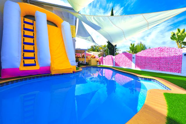 Bright backyard swimming pool with vivid blue water, a yellow-and-white inflatable water slide, pink floral privacy wall, triangular shade sails and green artificial turf — summer party setup