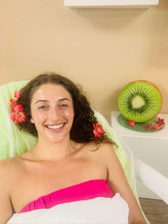 Relaxed woman smiling during a spa facial with glossy skin mask, pink towel, tropical flowers in her hair and a decorative kiwi slice on a side table — relaxing skincare treatment.