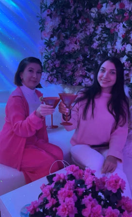 Two people clinking martini glasses in a pink floral indoor lounge with neon purple lighting and a table of bright pink blossoms