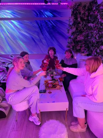 Five friends toasting pink cocktails over a charcuterie board in a cozy, galaxy-lit lounge with purple LED lighting and a floral wall