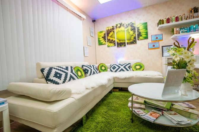 Bright, cozy spa waiting area with a white L-shaped sectional, kiwi-themed accent pillows and matching wall art, green shag rug, glass coffee table stacked with magazines and a floral vase.