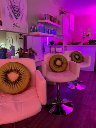 Magenta-lit beauty salon lounge with white swivel chairs featuring round kiwi-slice cushions, mirrored product shelves, decorative plants and modern wood flooring.