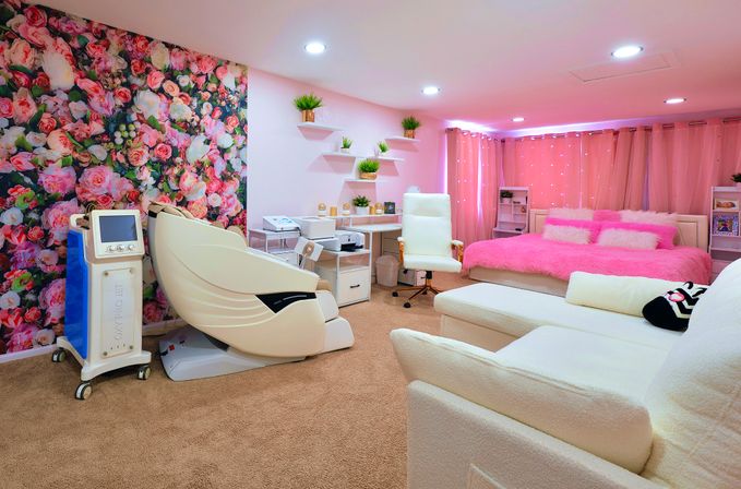 Cozy pink spa-style treatment room interior with a large floral accent wall, white massage pod chair, plush pink bed, white sofa and floating shelves with potted plants.