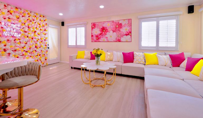 Bright modern lounge with U-shaped pale sofa, pink and yellow throw pillows, floral wall with neon "LOVE IS THE ANSWER", gold-accent coffee table, bar stool and pink flower artwork