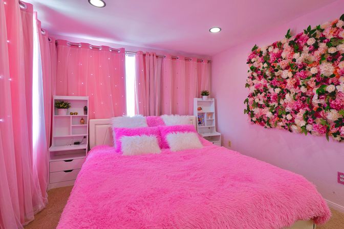 Dreamy pink bedroom with plush hot-pink faux-fur bedspread, white fluffy throw pillows, sheer pink curtains with tiny lights, white bedside shelving, and a large pink-and-white flower wall display.