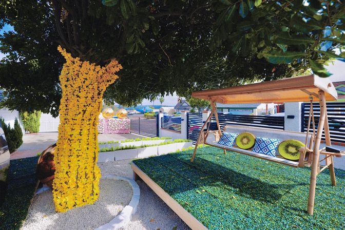 Sunny modern backyard patio with a wooden canopy swing on raised green turf, blue-and-white patterned and kiwi-print cushions, a large tree trunk wrapped in vibrant yellow flowers, pebble pathway and raised planters.