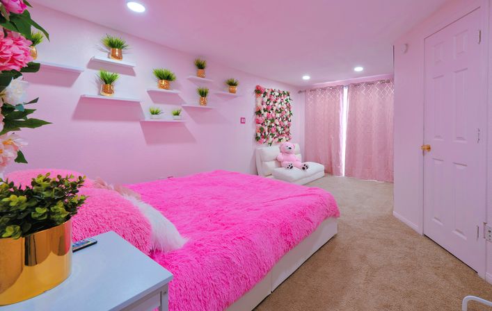 Cozy pink bedroom with a hot-pink faux-fur bedspread, white furniture, gold potted plants on floating shelves, a floral wall panel, and pink curtains.