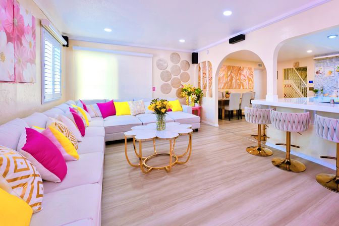 Bright, cheery open-concept living room and kitchen with large white L-shaped sofa, playful fuchsia and yellow throw pillows, marble nesting coffee table with gold base, tufted bar stools, light wood floors, arched doorways and floral wall art.