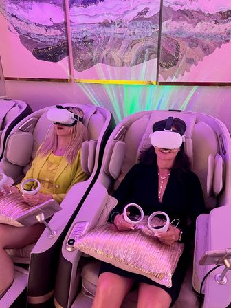Two women reclining in massage chairs wearing white VR headsets and holding controllers in a pink‑purple lit spa lounge, enjoying a virtual relaxation experience under abstract wall art and shimmering lights.