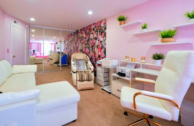 Cozy pink salon room with floral accent wall and mirrored closet doors, beige massage recliner, white office and sectional seating, and floating shelves with potted plants.