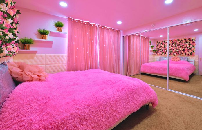 Vibrant pink bedroom with a fluffy hot-pink bedspread, floral wall panel, mirrored closet doors, sheer pink curtains with fairy lights, tufted headboard and floating shelves with potted greenery.