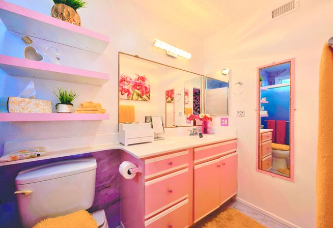 Cheerful pink bathroom with retro pink vanity and drawers, large wall mirror, open shelves, toilet, floral artwork and colorful accessories.