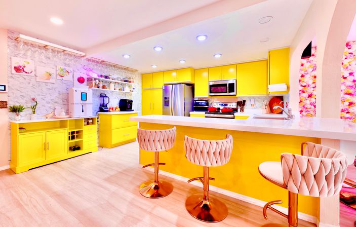 Cheerful modern kitchen with bright yellow cabinets and island, white quartz countertops, stainless steel appliances, three pink tufted swivel bar stools, light wood floors and a floral accent wall.