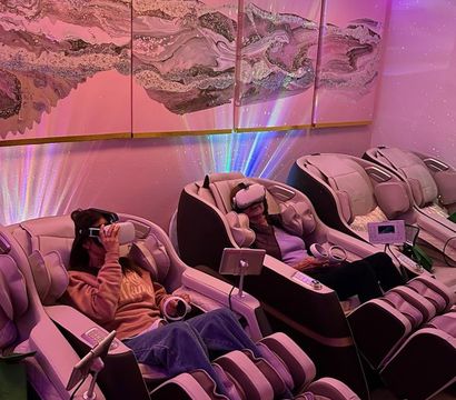 Cozy spa relaxation lounge with reclining massage chairs and VR headsets under pink-purple LED lighting, floral ceiling accents and abstract wall art.