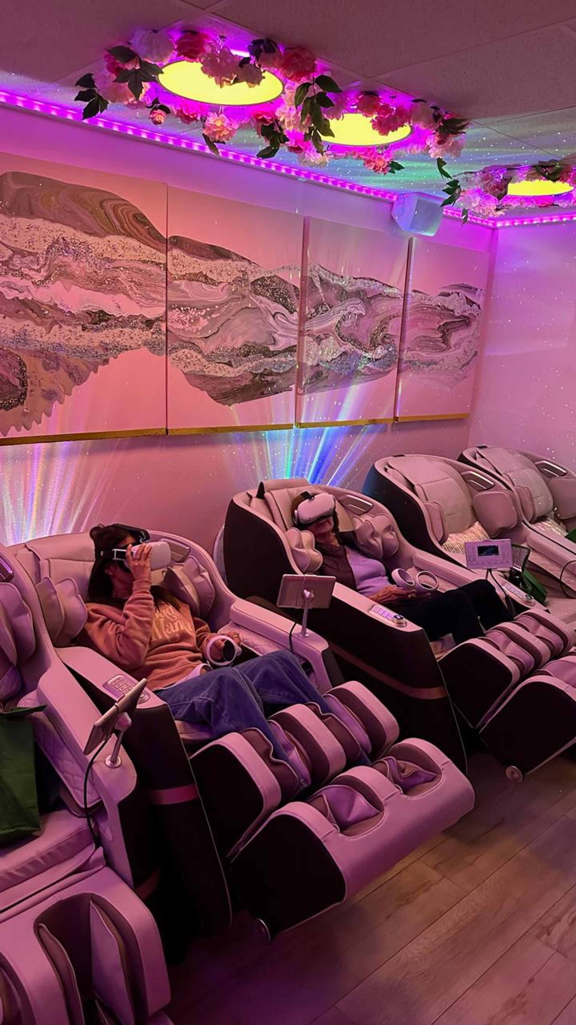 Cozy spa relaxation lounge with reclining massage chairs and VR headsets under pink-purple LED lighting, floral ceiling accents and abstract wall art.