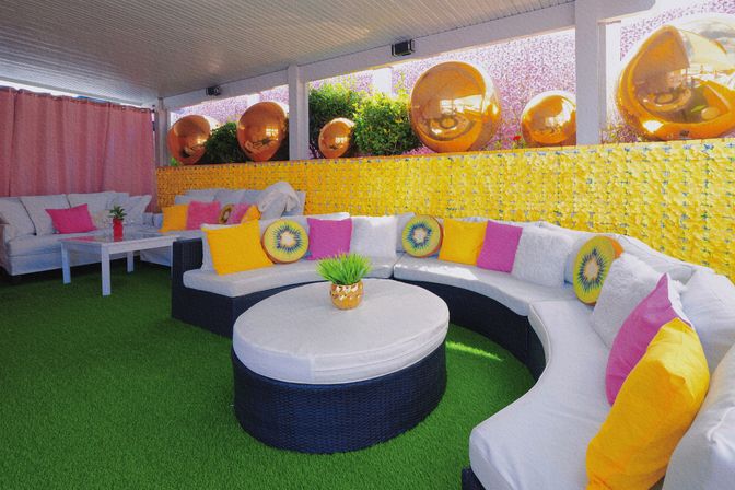 Covered outdoor patio lounge with curved wicker sectional and round ottoman on green artificial turf, bright yellow flower wall with reflective copper spheres, and colorful pink, yellow and kiwi-print throw pillows.