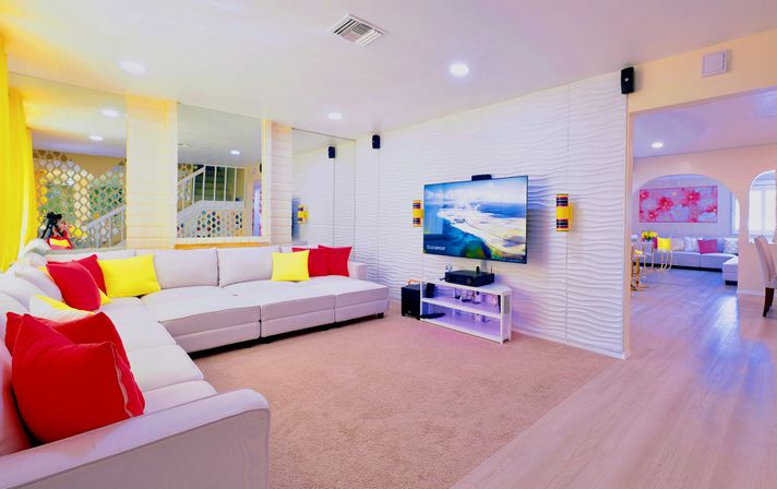 Bright modern open-plan living room with large white sectional, colorful red and yellow throw pillows, wall-mounted TV on a textured white accent wall, and archway to adjacent seating area.