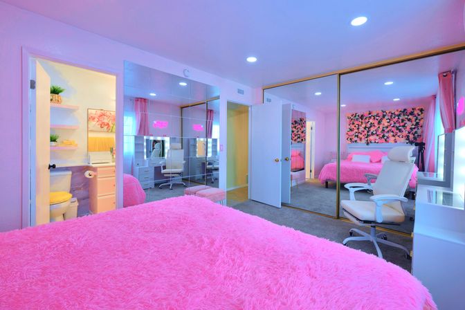 Vibrant pink bedroom with fluffy faux-fur bedspread, mirrored closet doors, floral accent wall, white vanity and office chair, and small ensuite bathroom