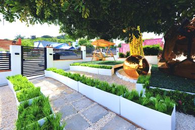 Modern suburban front yard with white raised planters of ornamental grass, stone-and-gravel pathway, black slatted gate, wooden swing under a tree, and a shiny golden sphere sculpture.