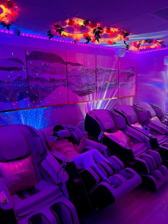 Purple LED-lit spa lounge with a row of reclining massage chairs, a person wearing a VR headset, floral ceiling rings and abstract wall art