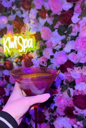 Hand holding red-stemmed martini glass with orange cocktail in front of a pink-and-purple illuminated flower wall and glowing neon sign