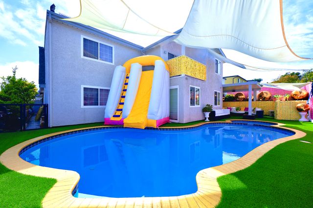 Vibrant backyard kidney‑shaped pool with a bright yellow inflatable slide attached to a two‑story stucco house, shaded lounge area, overhead sail canopies and artificial turf.
