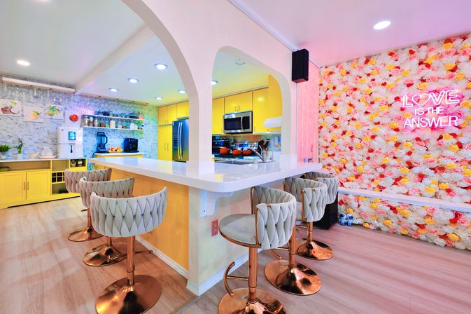 Bright Instagrammable café-style kitchen with yellow cabinets, white breakfast bar, quilted swivel stools on rose-gold bases, and a floral feature wall lit by a neon LOVE IS THE ANSWER sign.