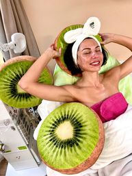 Smiling woman with bunny-ear headband relaxing during a spa facial, wrapped in a pink towel and surrounded by oversized kiwi-slice cushions in a bright wellness treatment room.