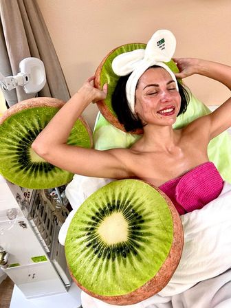 Smiling woman with bunny-ear headband relaxing during a spa facial, wrapped in a pink towel and surrounded by oversized kiwi-slice cushions in a bright wellness treatment room.