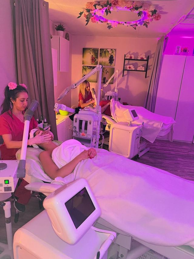 Neon-pink facial spa room with two estheticians performing skincare treatments on reclining beds surrounded by facial machines, towels, and floral ceiling decor.