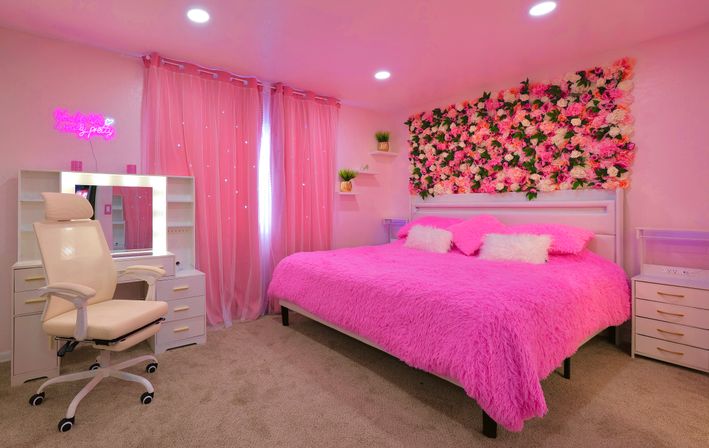 Instagram-ready pink bedroom with fuzzy hot-pink bedspread, floral rose wall backdrop, lighted vanity, sheer pink curtains, white swivel chair and potted plants.