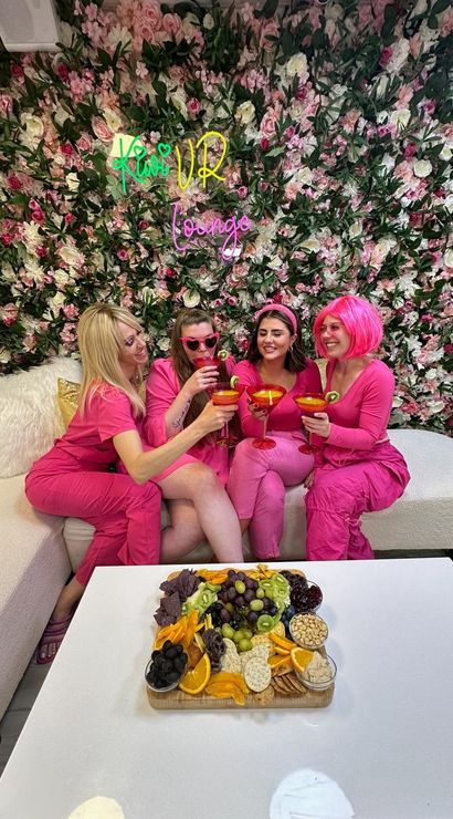 Four friends in vibrant pink outfits clinking kiwi-garnished cocktails on a sofa against a lush pink flower wall, with a colorful charcuterie board on the table.