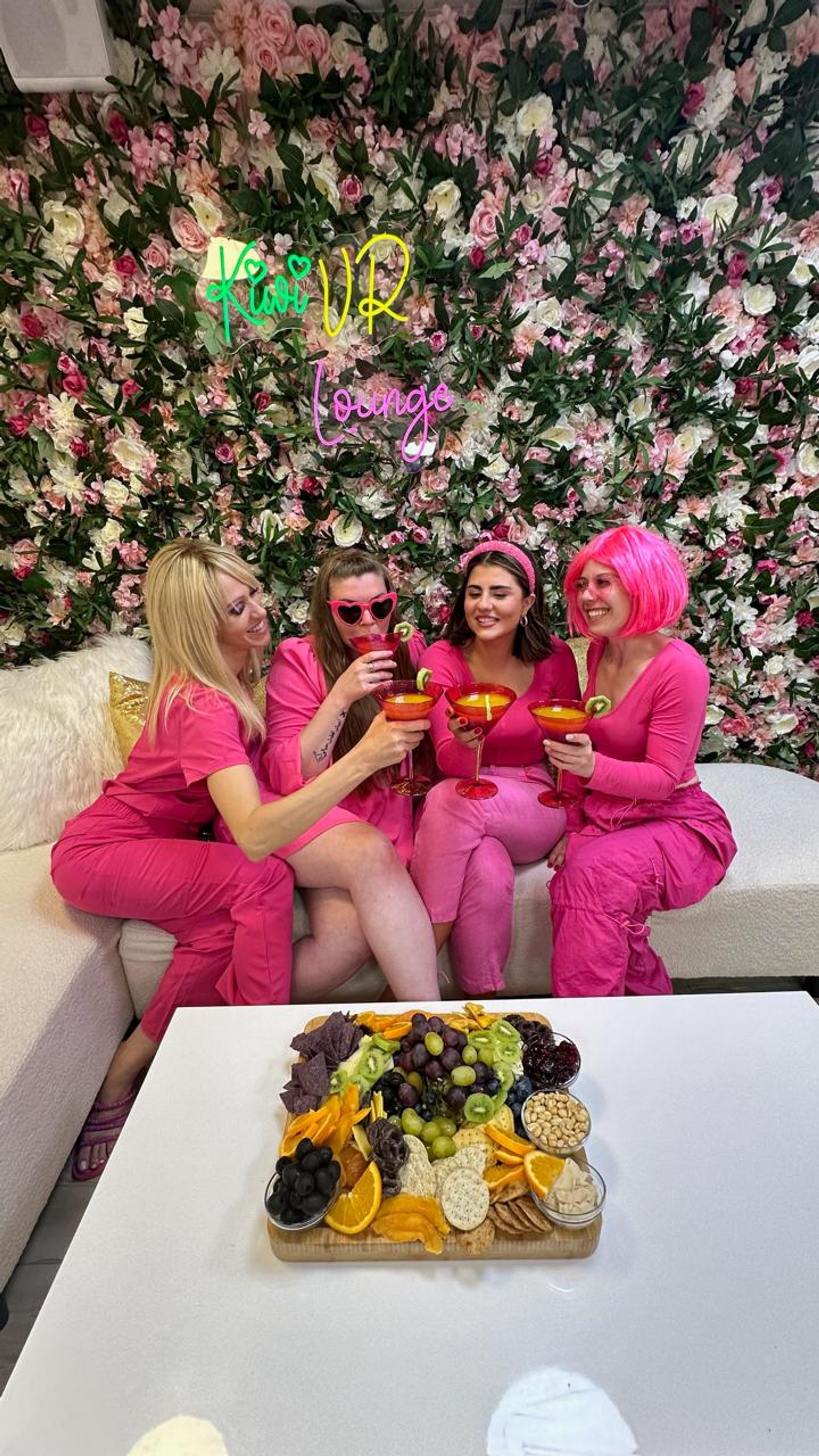 Four friends in vibrant pink outfits clinking kiwi-garnished cocktails on a sofa against a lush pink flower wall, with a colorful charcuterie board on the table.