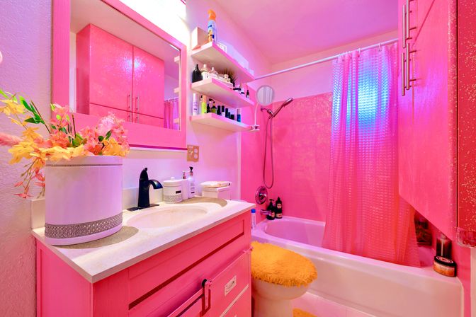 Vibrant pink home bathroom with pink vanity and mirror, floral arrangement on the sink, open shelves of toiletries, tub with pink shower curtain and yellow fuzzy toilet seat cover