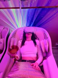 Person reclining in a pink-lit spa lounge massage chair wearing a VR headset and holding a cocktail glass beneath starry rainbow projector lights.