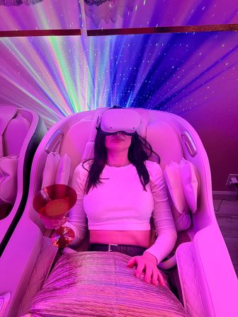 Person reclining in a pink-lit spa lounge massage chair wearing a VR headset and holding a cocktail glass beneath starry rainbow projector lights.